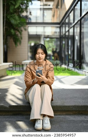 Portrait of pretty young Asian woman sitting front of business center and using mobile phone. Royalty-Free Stock Photo #2385954957