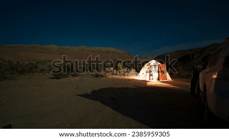 Camping on a bright starry night in the mountains. There is a white car, the light is on in the tent. Limestone mountains and canyons are visible in the distance. Green bushes grow. Sandy terrain.