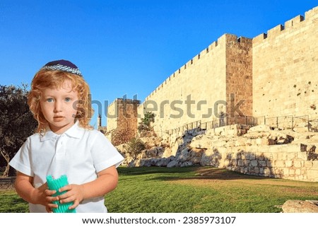 Charming blond little boy in a knitted kippah. Protective powerful walls of the Old City of Jerusalem at sunset. Jerusalem is the capital of Israel.  Royalty-Free Stock Photo #2385973107