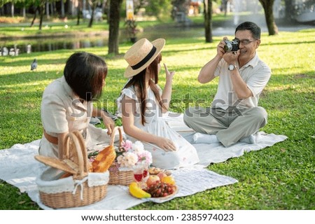 A happy Asian grandfather is taking a picture of his wife and granddaughter with his analog film camera while enjoying a picnic in the green park together. Happy family, family bonding