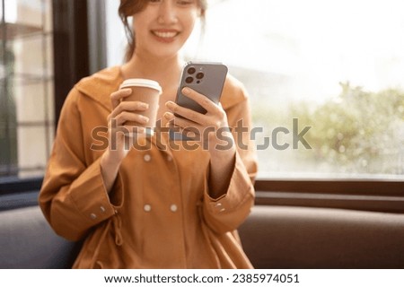 A beautiful and happy young Asian woman is enjoying her coffee and using her smartphone while relaxing in a coffee shop. Modern life concept