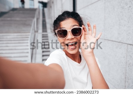 Young beautiful smiling hipster woman in trendy summer white t-shirt and jeans clothes. Carefree woman, posing in the street at sunny day. Positive model outdoors near wall. Takes Pov selfie 