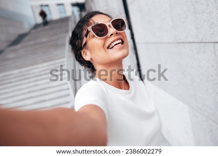 Young beautiful smiling hipster woman in trendy summer white t-shirt and jeans clothes. Carefree woman, posing in the street at sunny day. Positive model outdoors near wall. Takes Pov selfie 