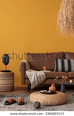 Creative composition of ethnic living room interior with brown sofa, yellow wall, braided pouf, patterned pillow, sculpture, grass lamp, patterned rug and personal accessories. Home decor. Template. Royalty-Free Stock Photo #2386003113