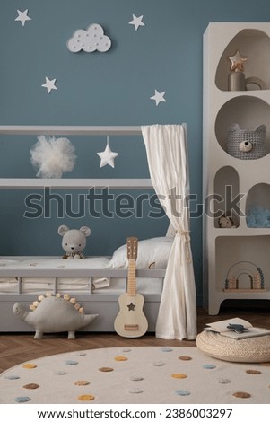 Aesthetic composition of cozy kid room interior with stylish bed, blue wall, braided pouf, round rug, books, colorful garland, gray curtain and personal accessories. Home decor. Template.