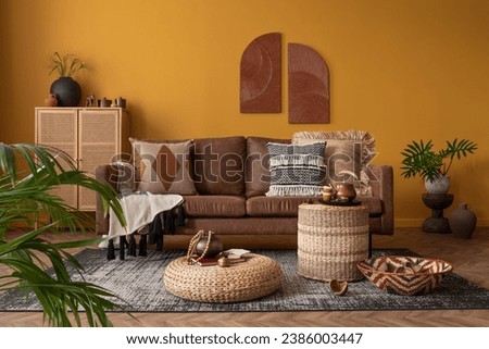 Warm and cozy composition of african living room interior with mock up poster frame, brown sofa, pouf, rattan sideboard, vase with leaves, pillows and personal accessories. Home decor. Template. Royalty-Free Stock Photo #2386003447