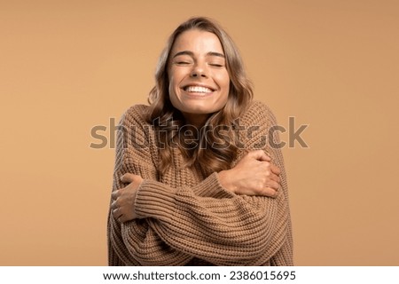 Happy smiling woman with closed eyes hugging herself wearing comfortable brown knitted sweater isolated on beige background. Winter concept Royalty-Free Stock Photo #2386015695