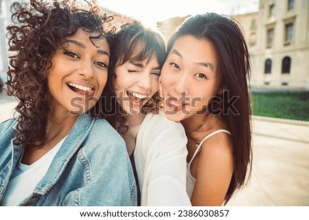 Three young women taking selfie pic with smart mobile phone device outside - Multiracial women smiling at camera together  Royalty-Free Stock Photo #2386030857