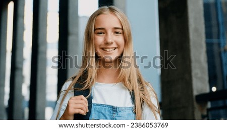 Portrait happy preteen girl holding book and looking at camera outdoors. Portrait of schoolgirl with backpack smiling at camera outside school building. Royalty-Free Stock Photo #2386053769