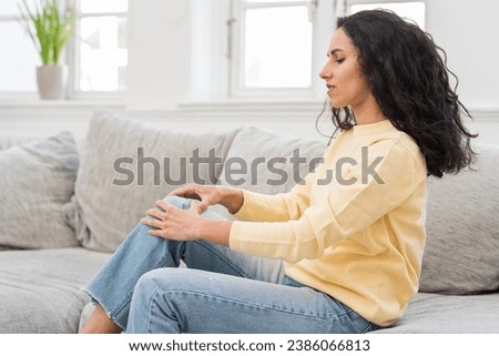 Side portrait of a young beautiful brunette worried about pain in her knee, holding her kneecap with her hands, massaging a bruise that hurts. Woman at home sitting on the sofa in the living room. Royalty-Free Stock Photo #2386066813