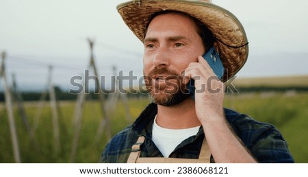 Close up portrait of happy young handsome man in hat having cellphone call. Farmer talking on smartphone standing in field in village. Organic smart farming and digital farming in the tea industry.