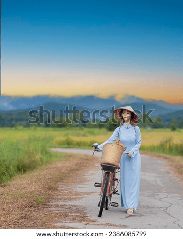 Beautiful Vietnamese Asian woman wearing a blue color Ao Dai National Costume Dress with red bicycle and flowers fresh yellow rice fields and mountain background. Portrait fashion show in nature.