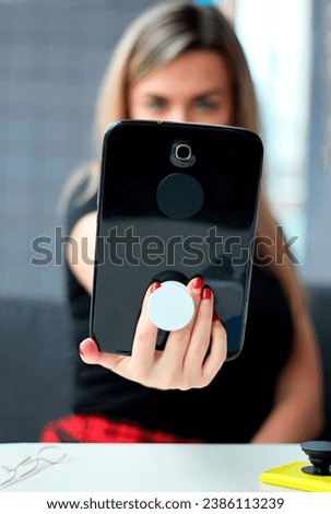 White blond girl holds a black tablet with the gadget holder (pop socket) and makes selfies. Woman is communicating online from a cafe or coworking space. Girl os on a video call.