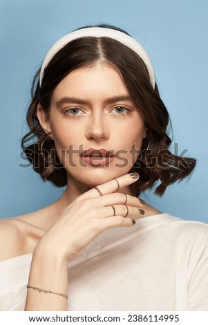 A brown-haired girl in a white blouse, rings, a bracelet and golden earrings is posing on the blue background. The girl's hair is fixed with a wide white headband covered with silky fabric.