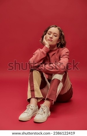 bored and stylish gen z girl in patchwork pants and cropped long sleeve sitting on red backdrop