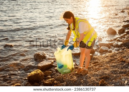 Clean up of pebble wild beach. Young volunteer Caucasian woman wearing vest and rubber gloves picking up trash. Concept of oceans pollution and garbage recycling.