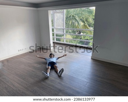 Photo of an happy little teeanger boy lies down on the floor on the back in an empty room from a modern appartement in a house wit a large window showing a jungle forest