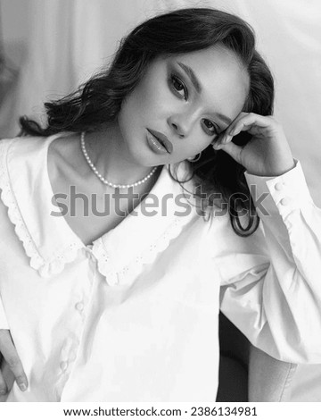 Black and white photo. Beautiful young girl in a white shirt and trousers on a white fabric background with a pearl necklace. Casual look. Lifestyle