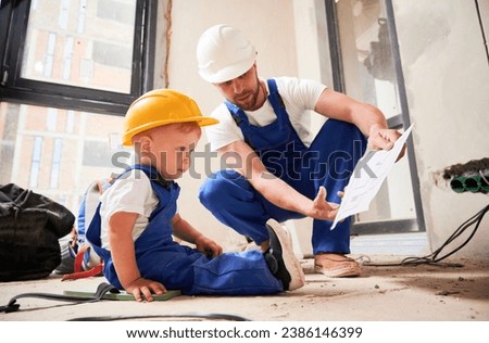 Male builder showing architectural drawings to little boy. Child in work overalls and construction helmet sitting on the floor and studying building plan with father in apartment under renovation. Royalty-Free Stock Photo #2386146399