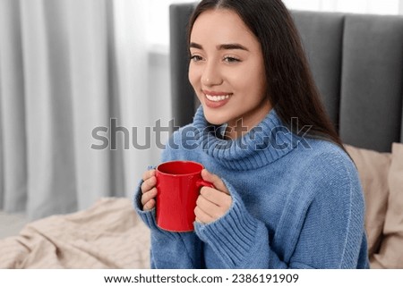 Happy young woman holding red ceramic mug on bed at home, space for text Royalty-Free Stock Photo #2386191909