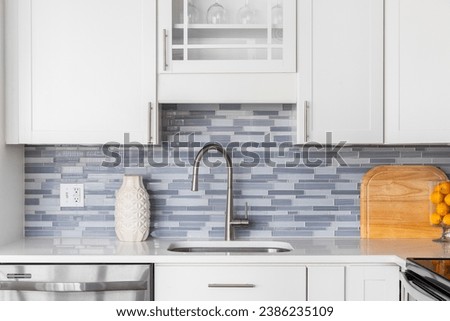 A kitchen faucet detail with a blue glass tile backsplash, white cabinets, decorations on the marble countertop, and a stainless faucet. Royalty-Free Stock Photo #2386235109