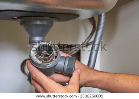 clogged sink pipe in hand,a plumber cleans a clogged siphon pipe of a sink in the kitchen Royalty-Free Stock Photo #2386250001