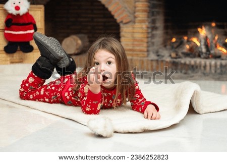 a little girl in winter pajamas smiles sweetly,lies comfortably on a warm woolen carpet near of a fire in the fireplace. Background For a project for the sale of warm winter accessories,clothes,shoes