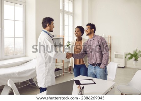 Smiling young multiracial couple handshake greeting with male doctor at consultation in clinic. Happy ethnic man and woman spouses shake hand make deal with GP in hospital. Healthcare concept. Royalty-Free Stock Photo #2386269045