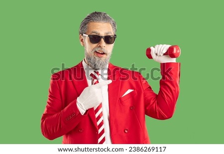 Grey haired old funny showman wearing red suit in Sants style and sunglasses holding red dumbbell in hand and pointing on it on green background. Christmas, New Year advertisement banner, marketing.