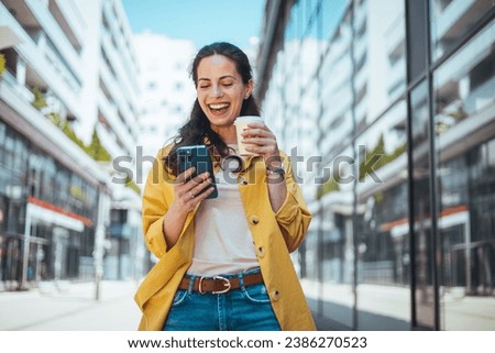 Modern young woman walking on the city street texting and holding cup of coffee. Business woman holding smartphone and looking away outdoors. Beautiful woman spending time in the city 
