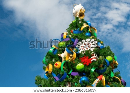 A tall outdoor Christmas tree with lush green fir branches, plastic ribbon circle garland, colorful balls, gold sheriff badge, snowflake and a plastic cartoon cat on the top holding a French horn. 