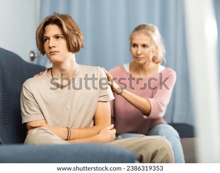 Portrait of upset and offended teenager sitting on sofa while mother soothing him. Mood swings and puberty concept Royalty-Free Stock Photo #2386319353