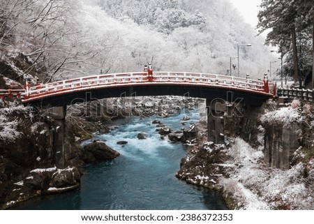 Shinkyo Sacred Bridge red, ancient architecture made from curved shaped wood spanning Daiya River, in winter covered by snow light snowfall. Tourist attraction and landmark in Nikko, Tochigi, Japan. Royalty-Free Stock Photo #2386372353