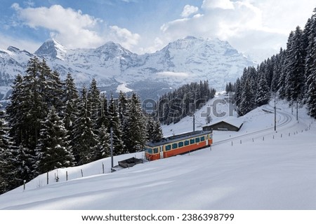 On a cold winter day, a local train makes its way thru forests and heavy snow on the hillside, with majestic Eiger, Monch and Jungfrau mountains in background, in Mürren, Berner Oberland, Switzerland