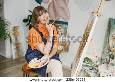 Elderly dreamful artist woman 50 years old wearing casual clothes sit near easel with painting artwork sit paint hold palette brush spend free spare time in living room indoor. Leisure hobby concept