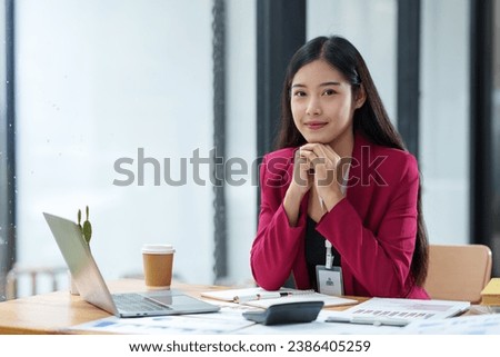 Confident Asian businesswoman smiling at camera while sitting at desk going through company financial business documents.