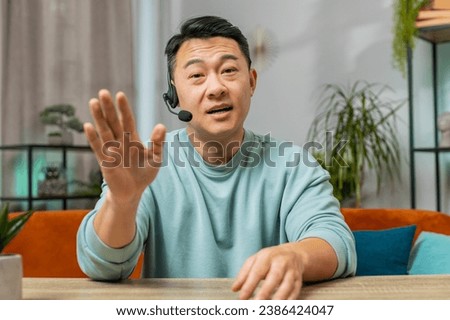 Asian Chinese man wearing headset, freelance worker call center or support service operator helpline. Chinese guy having talk with client or colleague communication support waving hello alone at home Royalty-Free Stock Photo #2386424047
