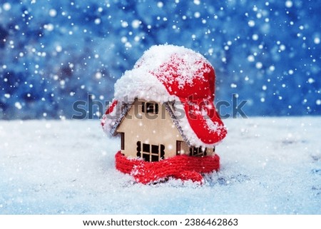 A wooden toy house in the snow in winter is covered with a hat and wrapped in a scarf. The concept of keeping warm Royalty-Free Stock Photo #2386462863