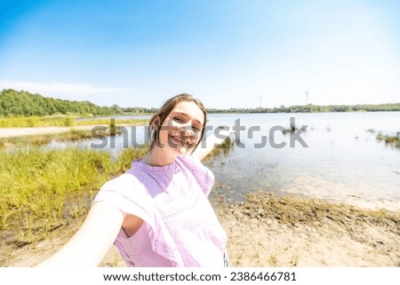 In the embrace of the great outdoors, a happy young woman snaps a selfie by the lakeside, her vibrant smile capturing the essence of a sun-kissed day. This snapshot is a celebration of health