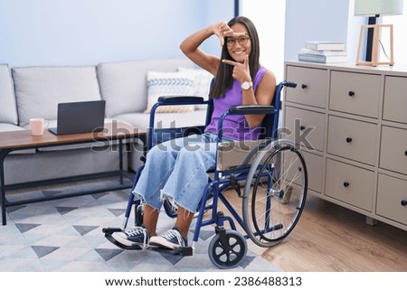 Young hispanic woman sitting on wheelchair at home smiling making frame with hands and fingers with happy face. creativity and photography concept. 