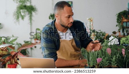 African american male inspection plants, greenhouse worker checking leaves and writing notes. Man small business owner florist working on desk in flower shop. Florist in apron looking at laptop making