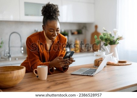 Smiling female freelancer scrolling social media on cellphone at desk in home office. Royalty-Free Stock Photo #2386528957
