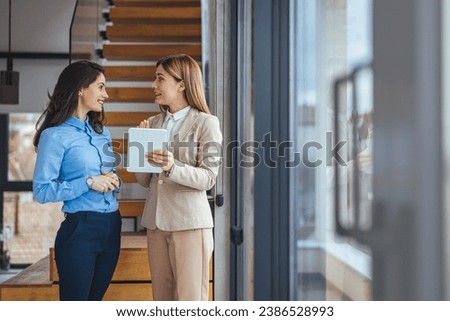 Two businesswomen chatting, meet together in hallway, share opinions and professional information, discuss project having business conversation or informal talk with tablet and coffee mug in office