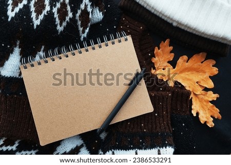 Notepad with black pen on a cozy sweater next to a hat and oak leaves