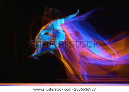 Young man, skateboarder with modern hairstyle practicing Ollie on skateboard with neon flashes of light isolated black background. Concept of youth culture, active lifestyle, style and fashion, art.