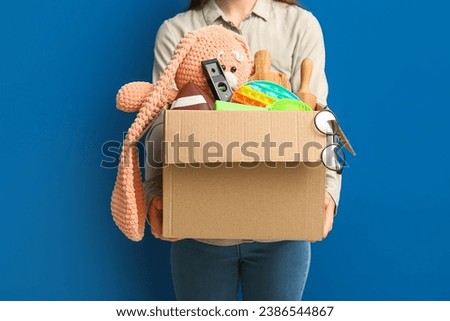 Woman holding box of unwanted stuff for yard sale on blue background Royalty-Free Stock Photo #2386544867