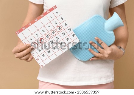 Young woman with hot water bottle and menstrual calendar on beige background, closeup Royalty-Free Stock Photo #2386559359