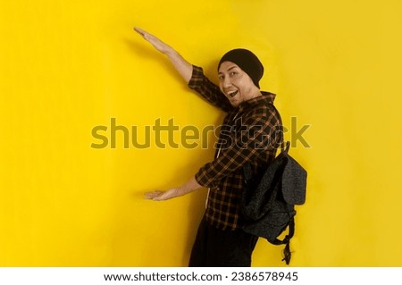 Cool funky young Asian man student with beanie hat and backpack presenting to the side for advertisement with excited expression Royalty-Free Stock Photo #2386578945