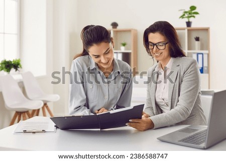 Business people working in modern office. Two young smiling happy female employees women sitting at the desk on workplace with financial documents, talking and going to sign a contract.