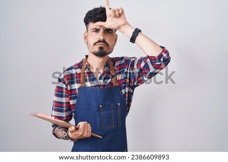 Young hispanic man with beard wearing waiter apron holding clipboard making fun of people with fingers on forehead doing loser gesture mocking and insulting. 
